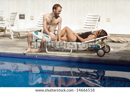 A young couple, share a pool chair, the woman lying, while the man sits near her feet. - horizontally framed