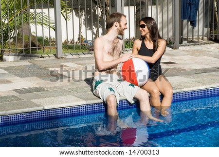 A man and a woman are relaxing together beside a pool.  They are looking at each other and dipping their legs in the water.  They are holding a beachball, Horizontally framed photo.