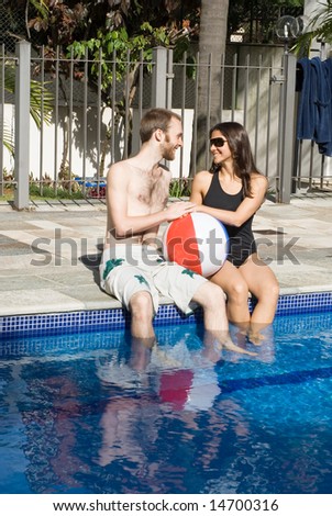 A man and a woman are relaxing together beside a pool.  They are looking at each other and dipping their legs in the water.  They are holding a beachball, Vertically framed photo.