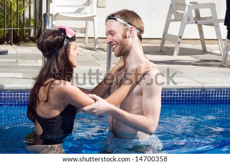 A man and woman are standing together in a pool.  They are hugging and looking at each other.    Horizontally framed photo.