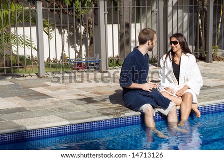 A man and a woman are relaxing together beside a pool.  They are looking at each other and dipping their legs in the water.  Horizontally framed photo.