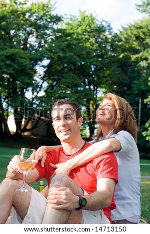 A couple sit in the grass, the man staring at the camera, the woman laughing. The man is drinking champagne while the woman is hugging the man from the back. - vertically framed