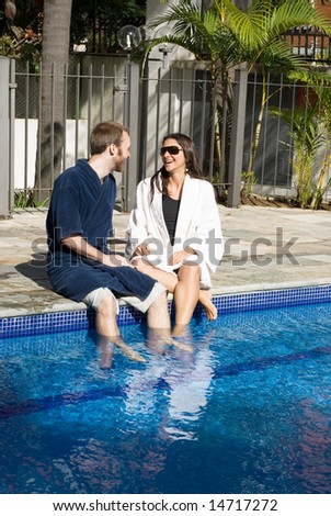 A man and a woman are relaxing together beside a pool.  They are looking at each other and dipping their legs in the water.  Vertically framed photo.