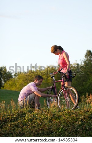 Woman holding on to her bike while the man crouches down to inspect it. Vertically framed photograph