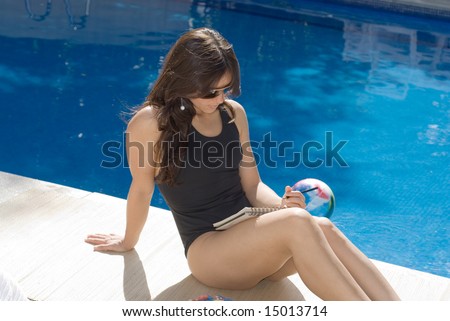 A young and attractive woman, sits down on the poolside floor, writing in a notepad. - horizontally framed
