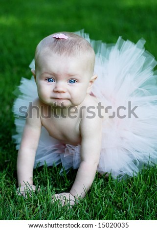 A baby, wearing a pink tutu, and a pink bow in her hair, crawls through the grass. Vertically framed shot.
