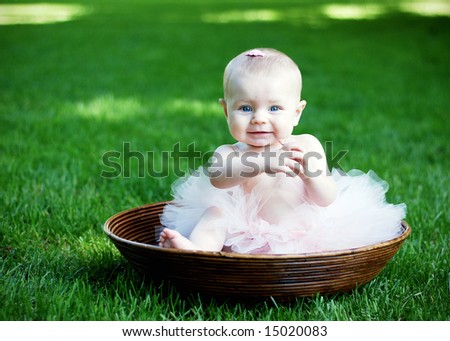 A young baby, wearing a pink tutu and bow in her hair, sits in a wooden bowl, staring at the camera. Horizontally framed shot.