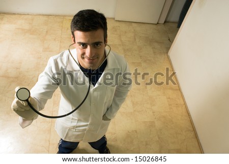 A young male doctor, stands, looking at the camera, holding his stethoscope. - horizontally framed