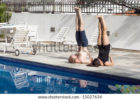 A man and a woman are exercising together beside a pool.  They are on the ground with their legs and torsos in the air.  They are looking away from camera.  Horizontally framed photo.