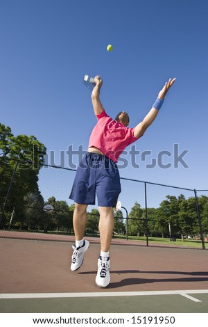 A man is outside on a tennis court playing tennis.  He is about to serve the ball and is looking away from the camera.  Vertically framed shot.