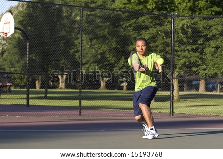 A man is outside on a tennis court playing tennis.  He is looking at the tennis ball.  Horizontally framed shot.