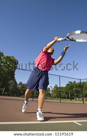 A man is outside on a tennis court playing tennis.  He had just served the ball and is looking at it in the air.  Vertically framed shot.