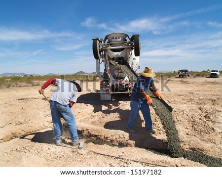 A cement truck is pouring cement into a foundation. A man is pointing the spout into a hole.  Horizontally framed shot.