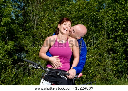 Woman laughs as man kisses her neck and hugs her while posing next to a mountain bike. Horizontally framed photograph.