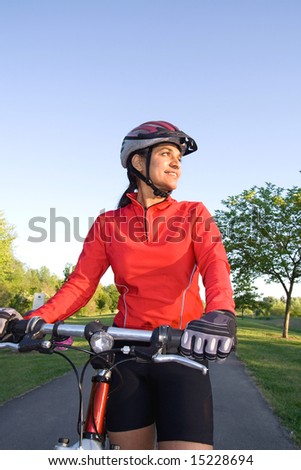 Close-up of woman standing next to bicycle and smiling in the park. Wearing sports gear and helmet. Vertically framed shot.