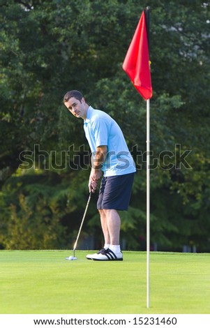 A man golfs while wearing a blue polo and blue pants. He stares at the hole. Vertically framed shot.