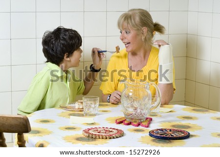Boy and woman sitting at table. Boy feeding woman. Woman wearing cast. Horizontally framed photo.
