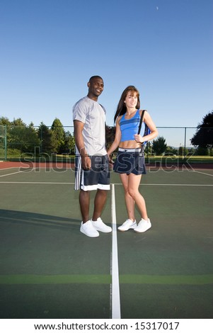A man and woman are standing together on a tennis court.  They are smiling and looking at the camera.  Vertically framed shot.