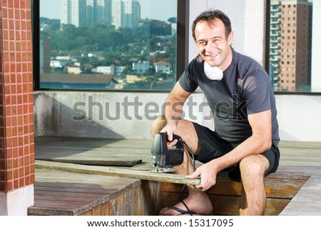 A man is standing in an unfinished apartment.  He is holding a power sander and smiling at the camera.  Horizontally framed shot.