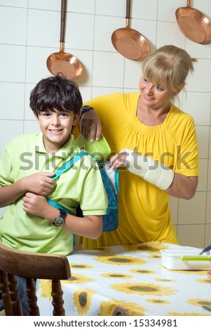 A mother is packing a lunch for her son in his backpack.  She is looking at him and he is looking at the camera.  Vertically framed shot.