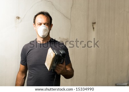 A man is standing in an unfinished apartment.  He is holding a power sander, wearing a mask, and looking at the camera.  Horizontally framed shot.