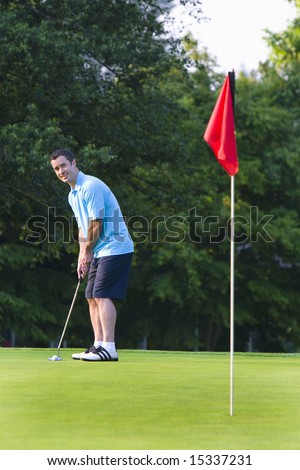 A man golfs while wearing a blue polo and blue pants. He smiles as he stares at the hole. Vertically framed shot.