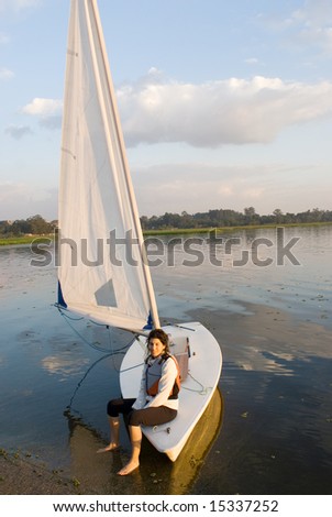 A woman smiling as she sits on the edge of the sailboat.  She is smiling, dangling her feet in the water, and looking away from the camera.  Vertically framed shot.