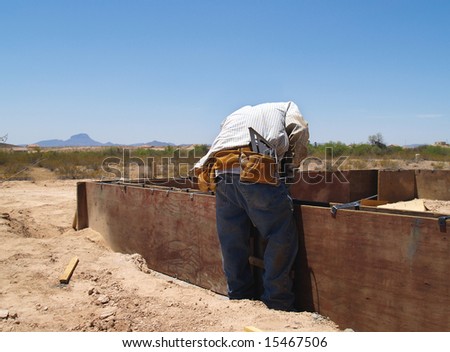 A construction worker is working on an excavation site.  He is bending down and looking at his work.  Horizontally framed shot.