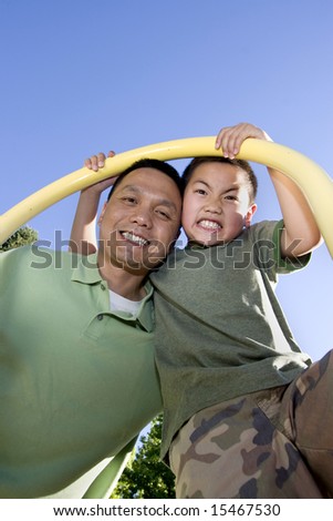 Father and son stand beneath fireman\'s pole on jungle gym. Father and son are looking down and smiling at the camera. Vertically framed photo.