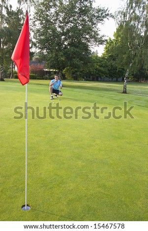 A young man, kneels, concentrating, attempting to line up his golf shot. Vertically framed shot.
