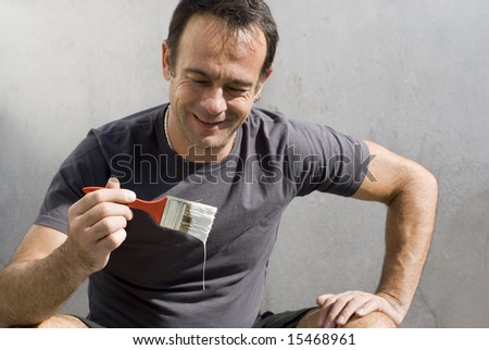 Man squatting smiling at paint dripping from paintbrush. He has hand on knee. Horizontally framed shot.