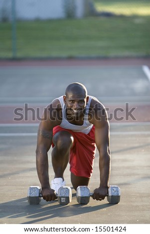 A fit man is crouching down on a tennis court.  He is holding weights in his hands.  He is smiling and looking at the camera.  Vertically framed shot.