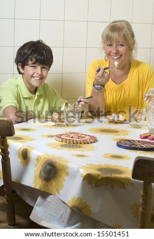 A woman and her child are sitting at a table together eating dinner.  They are both smiling and looking at the camera.  Vertically framed shot.