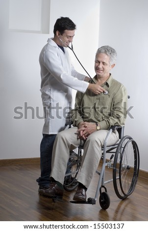 A patient is sitting in a wheelchair at a doctors office.  The doctor is listening to his heartbeat.  They are smiling and looking away from the camera.  Vertically framed shot.