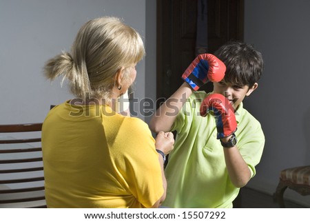 A woman and her son are playing and pretending to box each other.  He is wearing boxing gloves.  They are smiling and looking at the camera.  Horizontally framed shot.