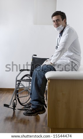 A young, male doctor is sitting on a bench in his office.  He is sitting next to a wheelchair.  He is smiling and looking at the camera.  Vertically framed shot.