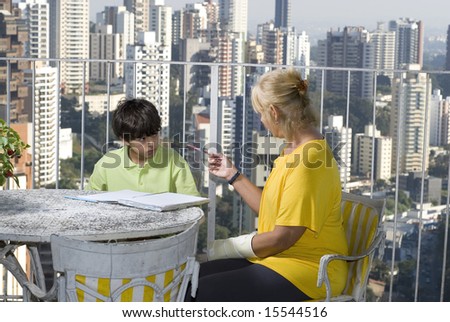 A woman and her child are sitting at a table.  She is instructing him out of a book.  They are looking at each other.  Horizontally framed shot.