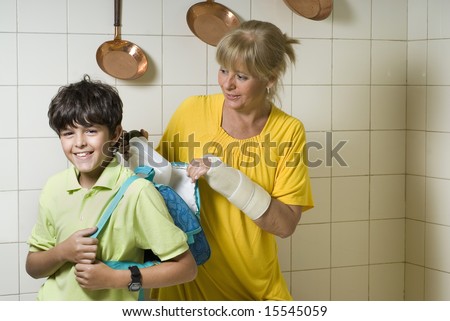 A mother is packing a lunch for her son in his backpack.  She is looking at him and he is looking at the camera.  Horizontally framed shot.