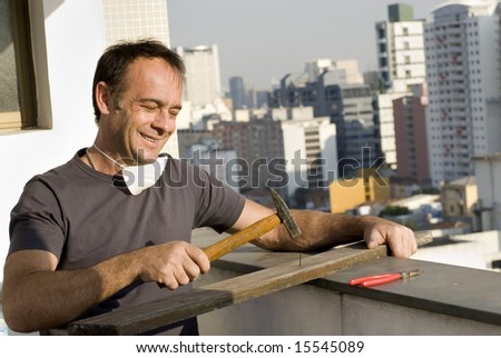 A man is about to pound a nail into a board with his hammer.  He is smiling and looking down at the nail.  Horizontally framed shot.