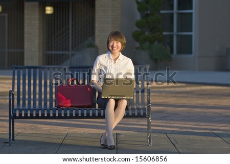 A woman smiling with her notebook PC on her lap, as she sits on a public bench. Horizontally framed shot.