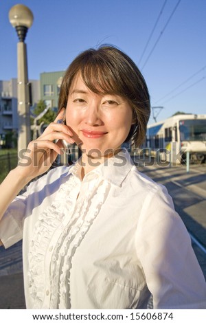 A woman stands smiling, talking on her cellular phone, standing in front of the bus station. Vertically framed shot.