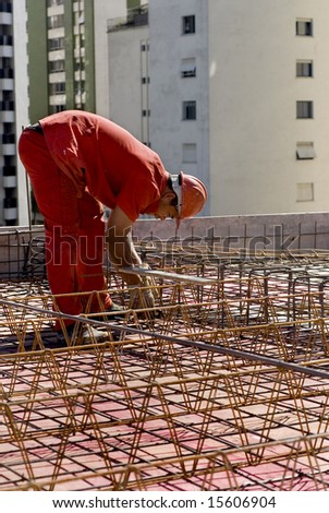 The worker is bent over the metal skeleton of the structure he is working on.  Buildings loom in the background over him.  Vertically framed shot.