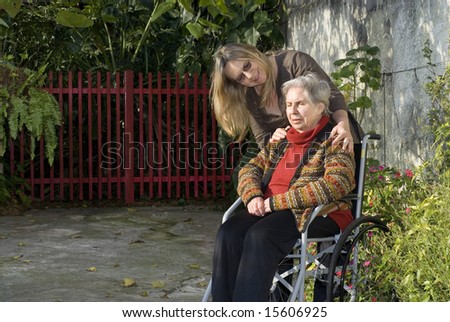A younger woman is with her elderly mother in a garden.  The older woman is sitting in a wheelchair.  She is smilng at her mother who is looking away.  Horizontally framed shot.
