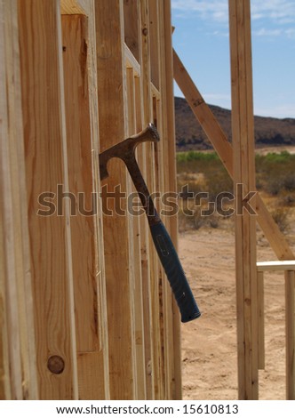 A house is being built on a construction site.  The walls have been put up and the frame can be seen.  A hammer is stuck into the wood of the frame.  Vertically framed shot.