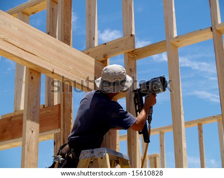 A man is  working on building a wall of a house.   He is on a ladder and his back is facing the camera.   Horizontally framed shot.