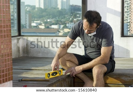 A man is sitting down using a spirit leveler for his construction job.  He is looking down at the leveler.  Horizontally framed shot.