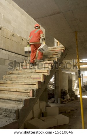 A construction worker is carrying heavy cinder blocks up the stairs at a construction site.  He has his back facing the camera.  Vertically framed shot.