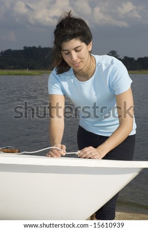 A woman is standing next to a sailboat on the water.  She is tying a knot in a rope, smiling, and looking down at the rope.  Vertically framed shot.