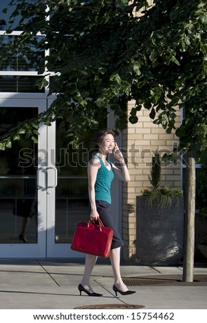 Young woman runs along building while talking on a cell phone. She is wearing a tank top and carrying a red bag. Vertically framed photo.