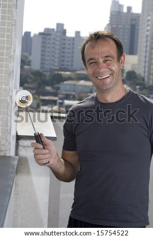A man is painting a wall in the city.  He is holding a paint roller, smiling, and looking at the camera.  Vertically framed shot.